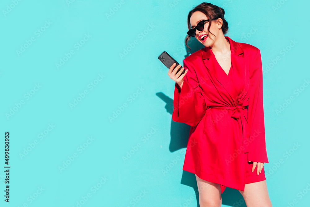 Young beautiful smiling female in trendy summer red dress. Sexy carefree woman posing near blue wall in studio. Positive model having fun.Looking at smartphone screen and using apps. In sunglasses Young beautiful smiling female in trendy summer red dress. Sexy carefree woman posing near blue wall in studio. Positive model having fun.Looking at smartphone screen and using apps. In sunglasses