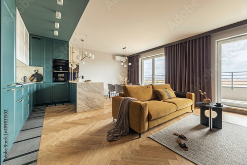 Creative and modern vintage open space interior design with yellow sofa and turqoise kithen with a marble island. Grey carpet on the herringbone parquet with coffe table, brown curtain in windows. 
 photo