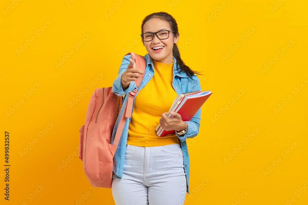 Portrait of excited young Asian woman student in denim clothes with backpack holding notebook, pointing fingers at camera on yellow background. Education in high school university college concept Portrait of excited young Asian woman student in denim clothes with backpack holding notebook, pointing fingers at camera on yellow background. Education in high school university college concept