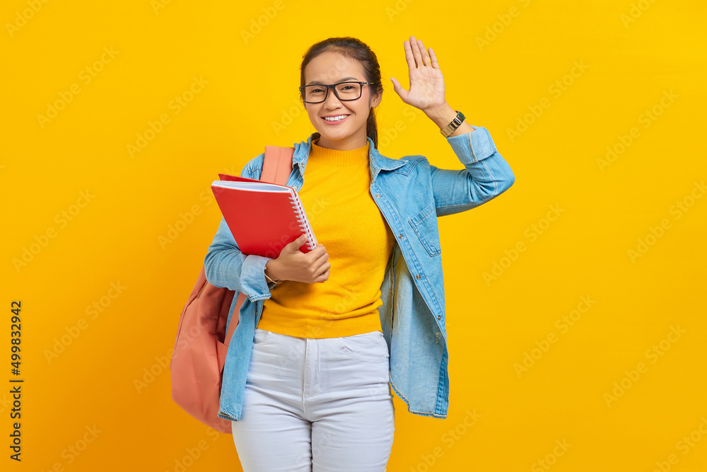 Smiling young Asian woman student in denim clothes with backpack holding notebook, waving and greeting with hand as notices someone isolated on yellow background Smiling young Asian woman student in denim clothes with backpack holding notebook, waving and greeting with hand as notices someone isolated on yellow background