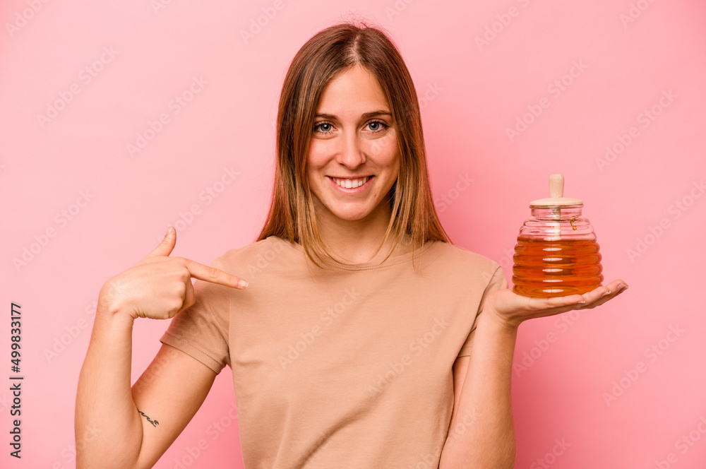 Fototapeta premium Young caucasian woman holding honey isolated on pink background person pointing by hand to a shirt copy space, proud and confident