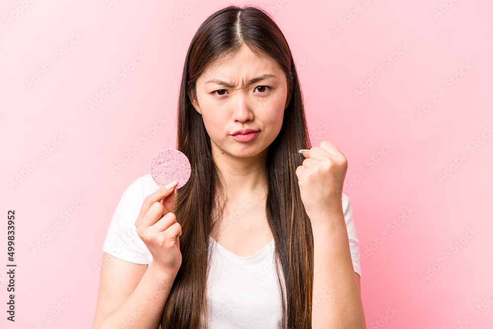 Young asian woman holding facial sponge isolated on pink background showing fist to camera, aggressive facial expression. Young asian woman holding facial sponge isolated on pink background showing fist to camera, aggressive facial expression.