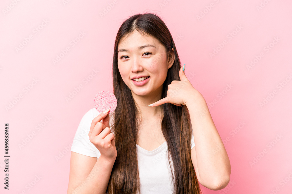 Young asian woman holding facial sponge isolated on pink background showing a mobile phone call gesture with fingers. Young asian woman holding facial sponge isolated on pink background showing a mobile phone call gesture with fingers.
