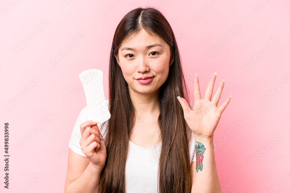 Young asian woman holding sanitary napkin isolated on pink background smiling cheerful showing number five with fingers. Young asian woman holding sanitary napkin isolated on pink background smiling cheerful showing number five with fingers.