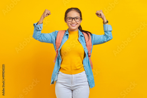 Cheerful young Asian woman student in denim outfit with backpack raises arms and shows biceps isolated on yellow background. Education in university concept