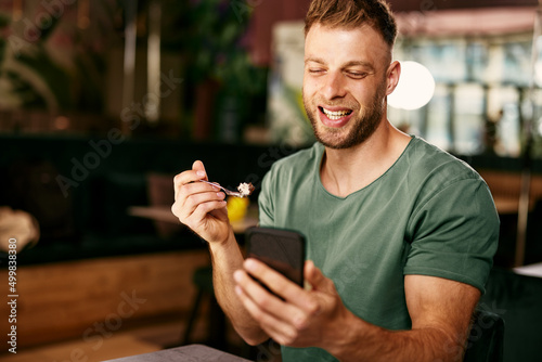 Handsome young man eating dessert in a cafe and uses a smartphone