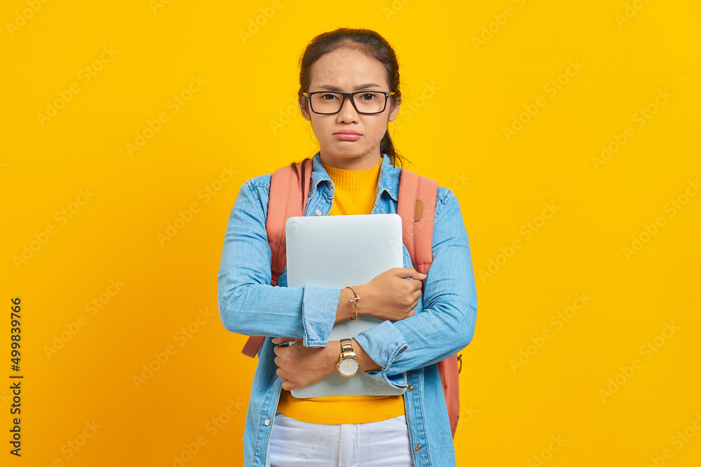 Portrait of displeased young Asian woman student in casual clothes with backpack holding laptop and looking at camera isolated on yellow background. Education in university college concept Portrait of displeased young Asian woman student in casual clothes with backpack holding laptop and looking at camera isolated on yellow background. Education in university college concept