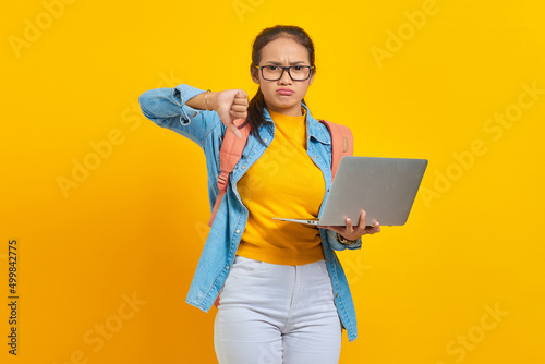 Portrait of angry young Asian woman student in casual clothes with backpack using laptop and showing thumbs down with finger isolated on yellow background. Education in university college concept