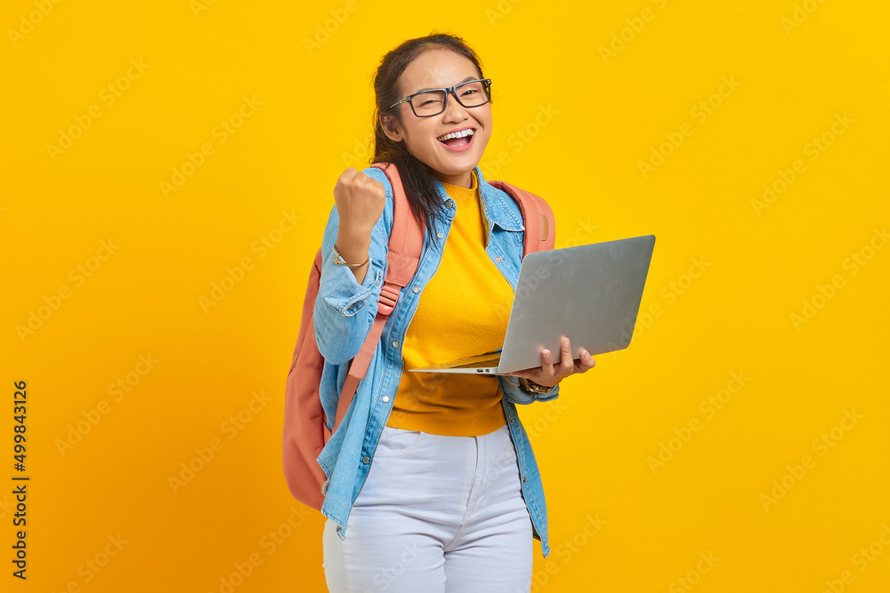 Portrait of excited young Asian woman student in casual clothes with backpack using laptop and celebrating success isolated on yellow background. Education in university college concept