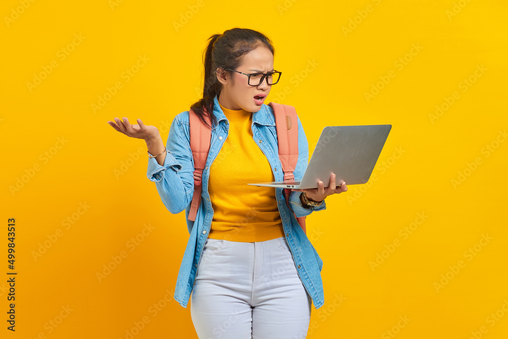 Portrait of annoyed young Asian woman student in casual clothes with backpack showing copy space on palm and holding laptop isolated on yellow background. Education in university college concept