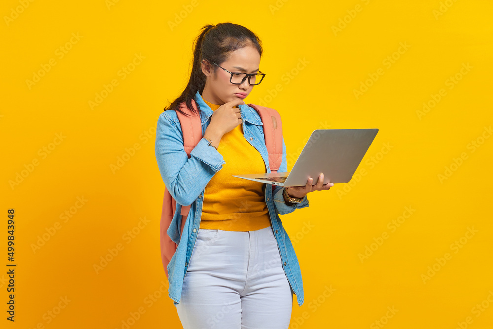 Portrait of serious young Asian woman student in casual clothes with backpack using laptop and thinking about question isolated on yellow background. Education in university college concept Portrait of serious young Asian woman student in casual clothes with backpack using laptop and thinking about question isolated on yellow background. Education in university college concept