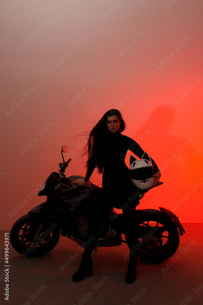 Fototapeta premium young teen brunette girl with long legs stands fashion with a biker helmet with ears near black sports motorcycle on a white wall background in red studio light. lifestyle fashion concept, free space