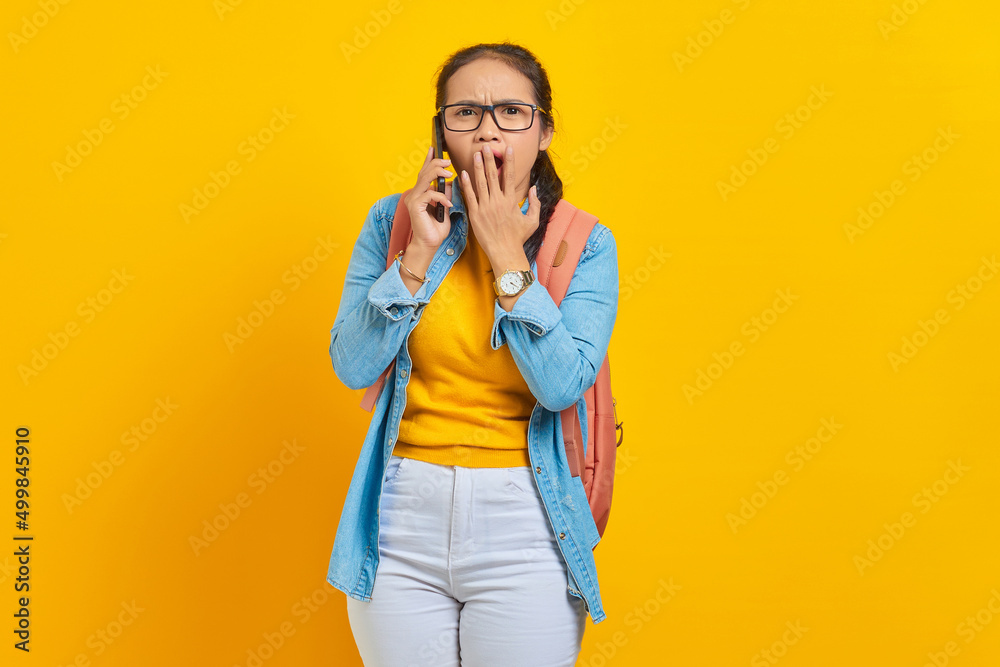 Portrait of surprised young Asian woman student in casual clothes with backpack holding smartphone and covering mouth with palms isolated on yellow background. Education in college university concept Portrait of surprised young Asian woman student in casual clothes with backpack holding smartphone and covering mouth with palms isolated on yellow background. Education in college university concept