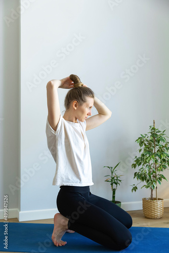 caucasian woman is doing hair bun, having a break during sport exercise, sitting on blue matt, at her home, flowers pot, white background