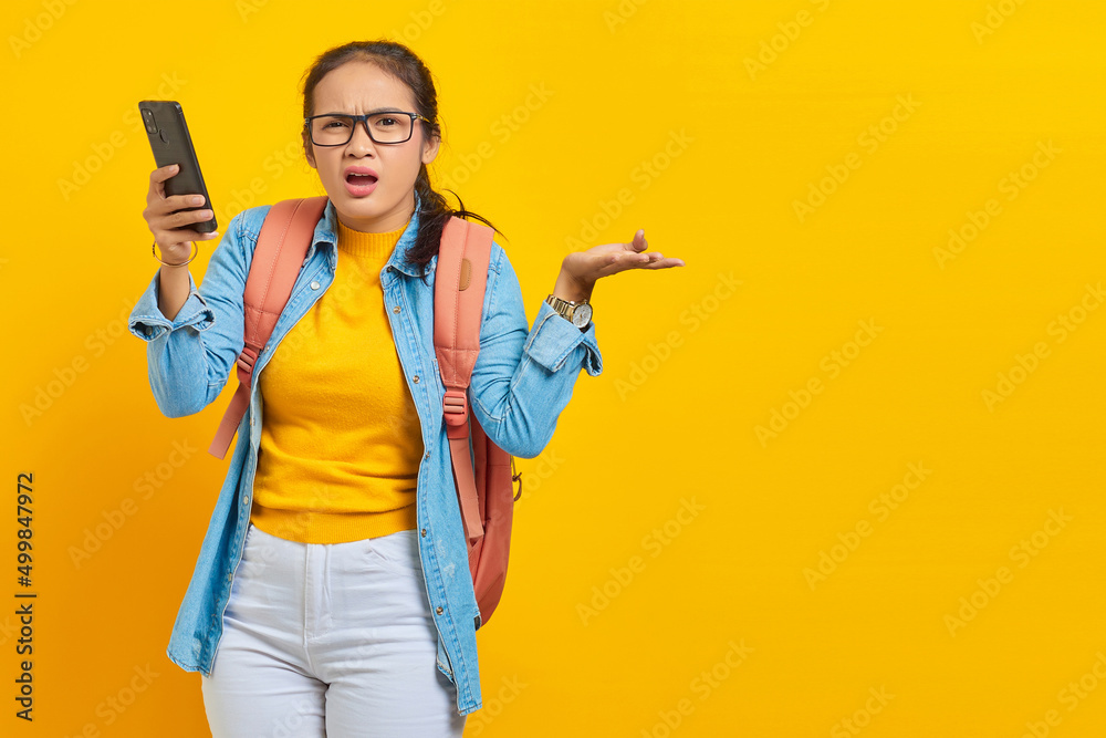 Portrait of confused young Asian woman student in casual clothes with backpack holding mobile phone and saying no with palms isolated on yellow background. Education in college university concept Portrait of confused young Asian woman student in casual clothes with backpack holding mobile phone and saying no with palms isolated on yellow background. Education in college university concept