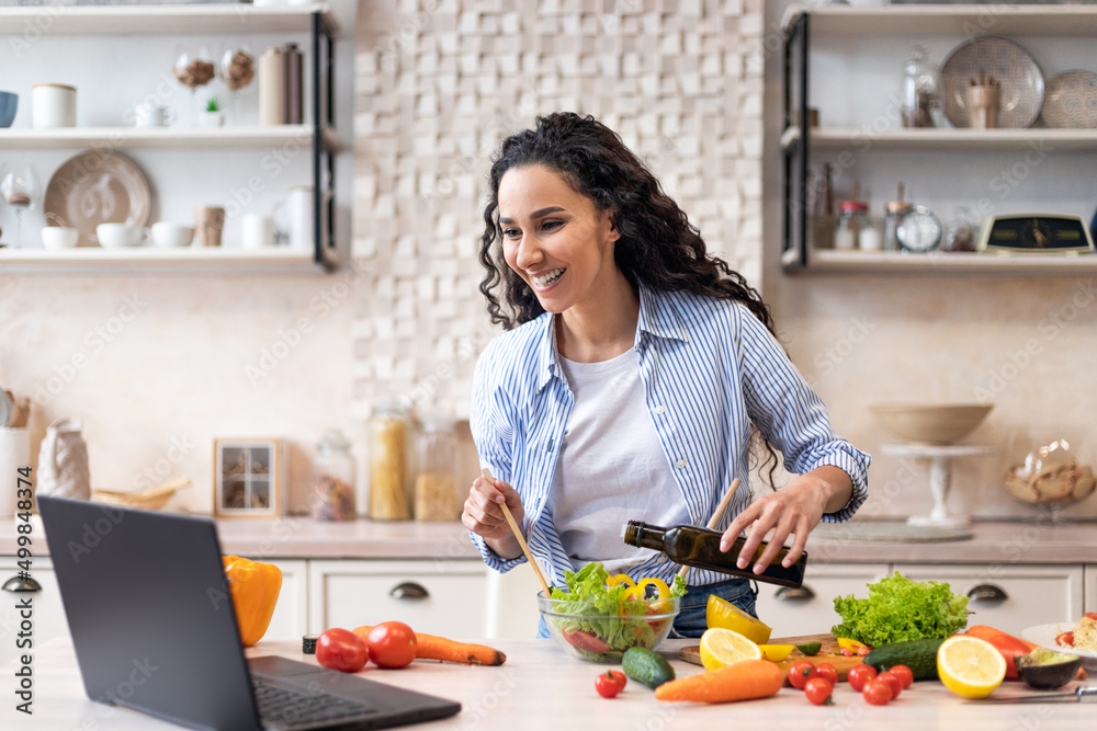 Happy lady watching video recipe on laptop while cooking healthy lunch in kitchen, using computer
