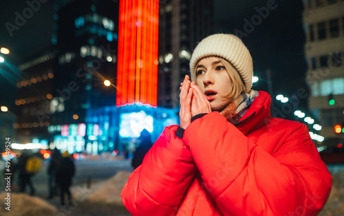 Beautiful woman warms her hands by breathing on them outdoors in sub-zero winter temperatures against the backdrop of a night landscape with modern illuminated architecture