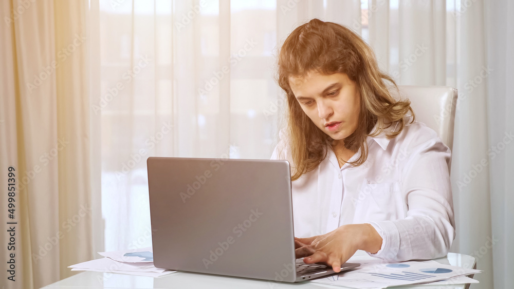Disabled woman freelancer with cerebral palsy works online on laptop from home office sitting at table against window behind curtain in room. Disabled woman freelancer with cerebral palsy works online on laptop from home office sitting at table against window behind curtain in room.