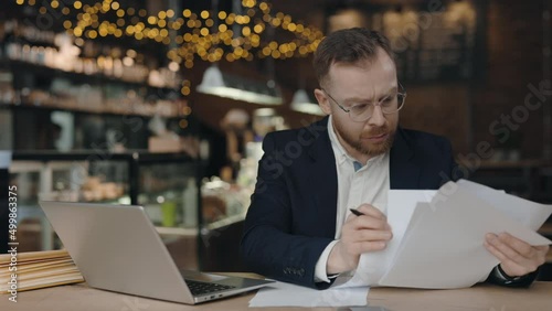 Clever and concentrated businessman doing paperwork. Man looking through work paper while sitting at the table with laptop at the cafe. Man checking report and collate data concept