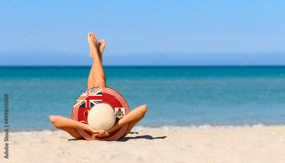 Fototapeta premium A slender girl on the beach in a straw hat in the colors of the Bermuda flag. The concept of a perfect vacation in a resort in the Bermuda. Focus on the hat.