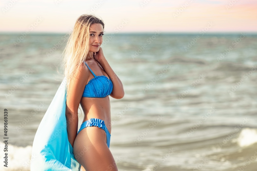 A girl with blond hair in a bluish swimsuit and a bright shawl walks along the beach