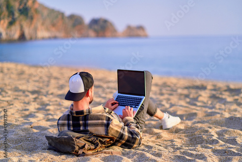 Millennial freelancer guy using mockup laptop with empty screen blank and lying on the sand beach by the sea. Dream office remote work concept