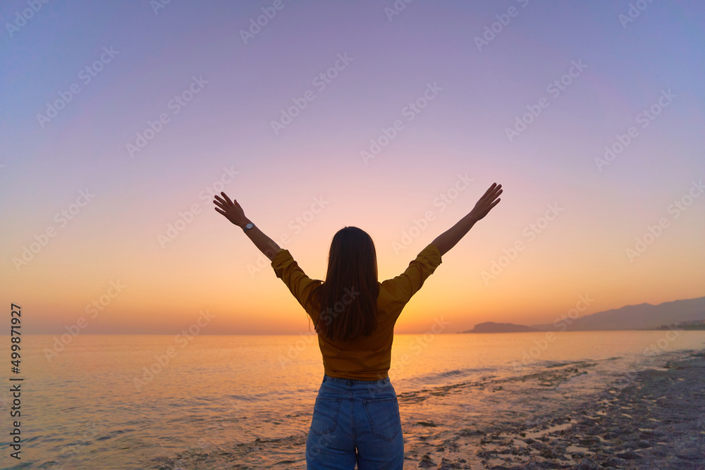 Back view of free calm bliss satisfied woman standing with open arms against of sunset sky by the sea in a happy beautiful inspired moment of her life