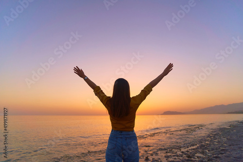 Back view of free calm bliss satisfied woman standing with open arms against of sunset sky by the sea in a happy beautiful inspired moment of her life