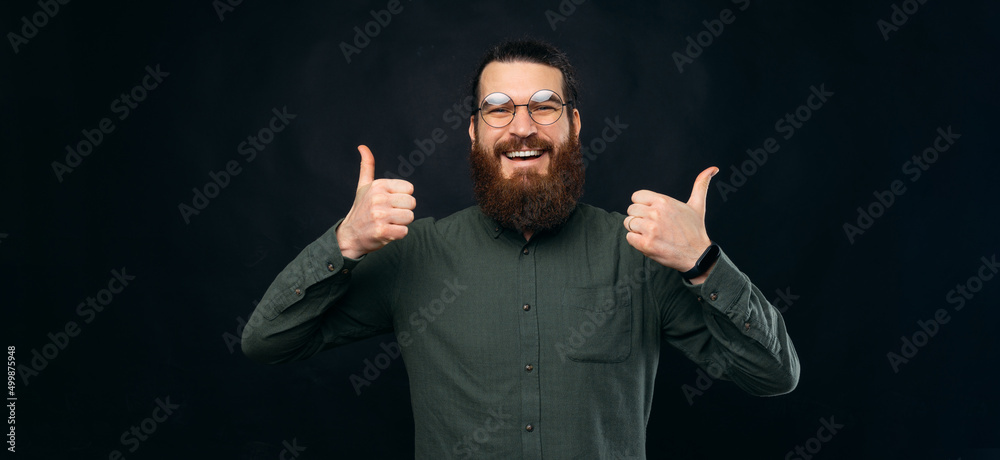 Panorama size photo of a young ecstatic bearded man showing thumbs up. Studio shot over black backdrop. Panorama size photo of a young ecstatic bearded man showing thumbs up. Studio shot over black backdrop.