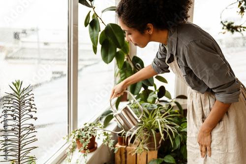 Foto Young black woman wearing apron watering plants in home