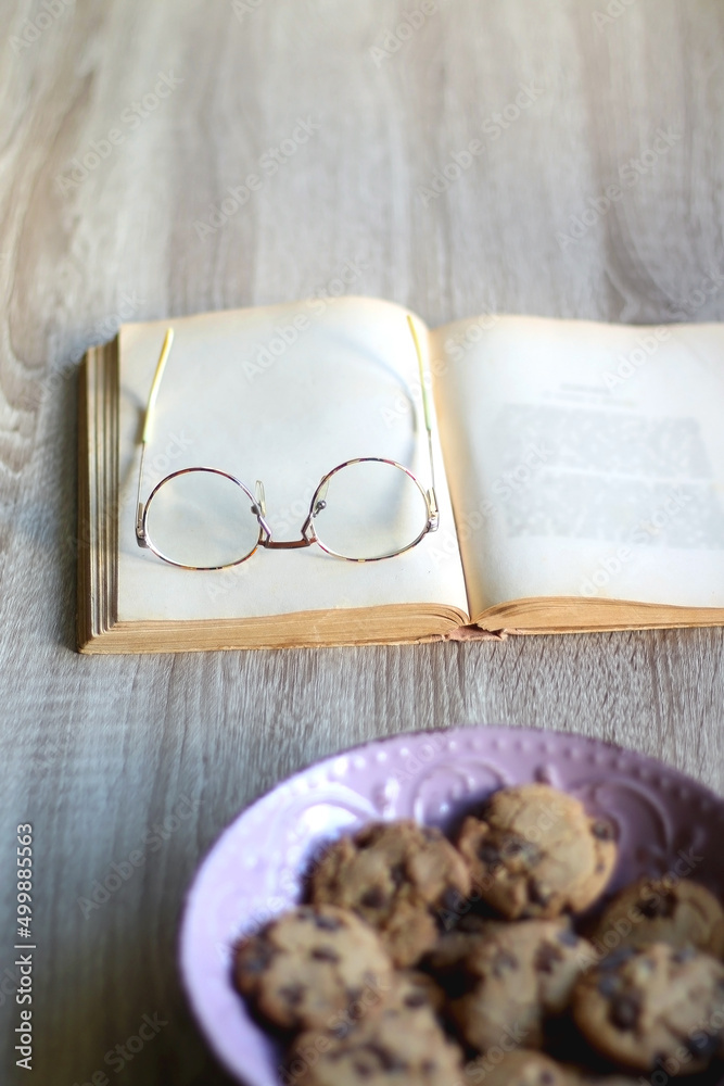 Fototapeta premium Bowl of chocolate chip cookies, open book and reading glasses on the table. Selective focus.