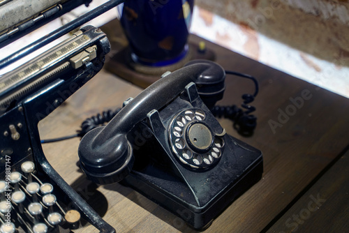 Old dust-covered black phone with disk dialing on a wooden table next to the typewriter