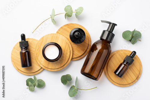 Natural cosmetics concept. Top view photo of glass bottles and cream jar on wooden stands and eucalyptus leaves on isolated white background photo