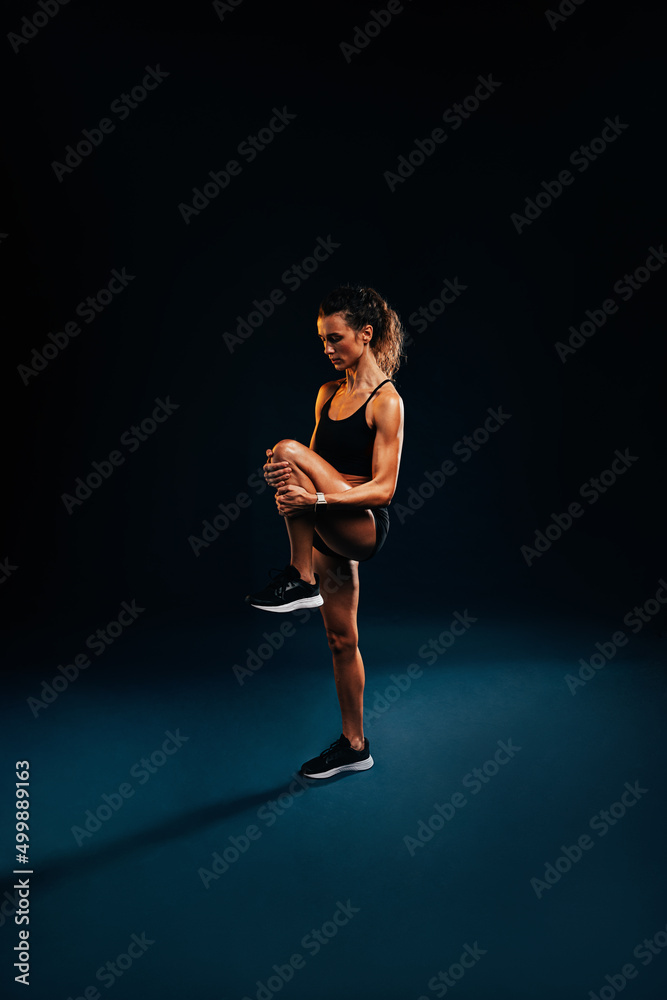 Fitness woman stretching her leg against dark background. Female runner doing stretches in studio.