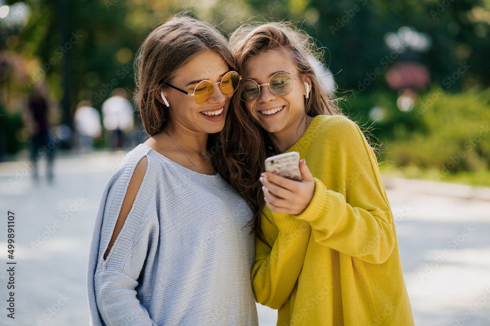 Fototapeta premium Girls outside in bright clothes and sunglasses in headphones, holding smartphone. Video call, online time with friends, outdoor time, warm sunny day in city