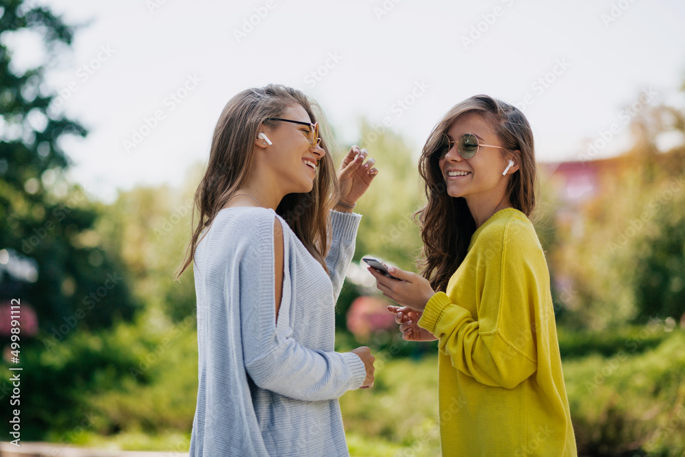 Pretty modern women with long wavy hairstyle in bright clothes are talking and listening music in sunny warm day on city background Pretty modern women with long wavy hairstyle in bright clothes are talking and listening music in sunny warm day on city background
