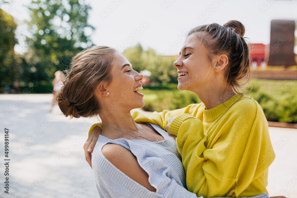 Two emotional female friends wearing in yellow and blue sweaters is laughing and hugging while walking in the park