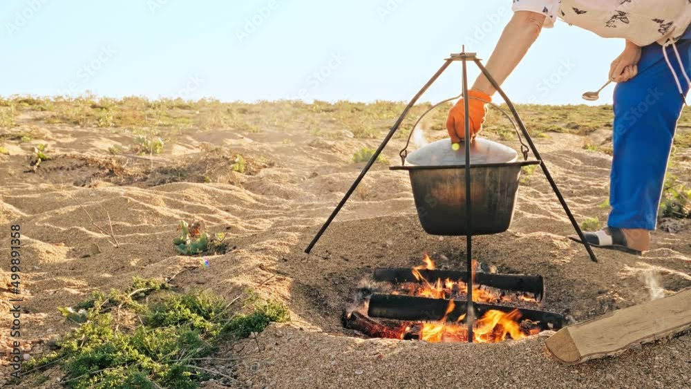 Campground chef opens the metal lid of the pot and starts stirring the food in the cauldron. Steam from the pot flutters in the wind in slow motion. Concept of simple life by the sea under blue sky. Campground chef opens the metal lid of the pot and starts stirring the food in the cauldron. Steam from the pot flutters in the wind in slow motion. Concept of simple life by the sea under blue sky.