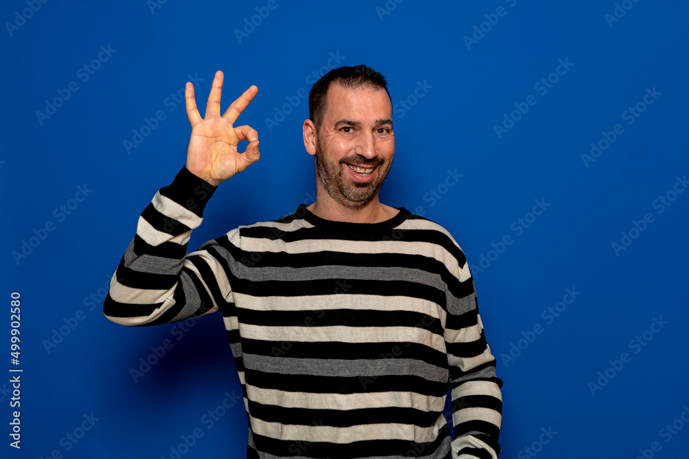 Naklejka premium Spanish bearded man in his 40s wearing a striped pullover making the ok gesture while smiling, isolated over a blue studio background