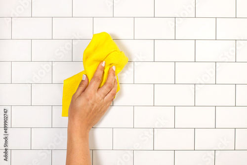 Mano de mujer limpiando con un paño amarillo una pared con azulejos. Vista de frente y de cerca. Copy space