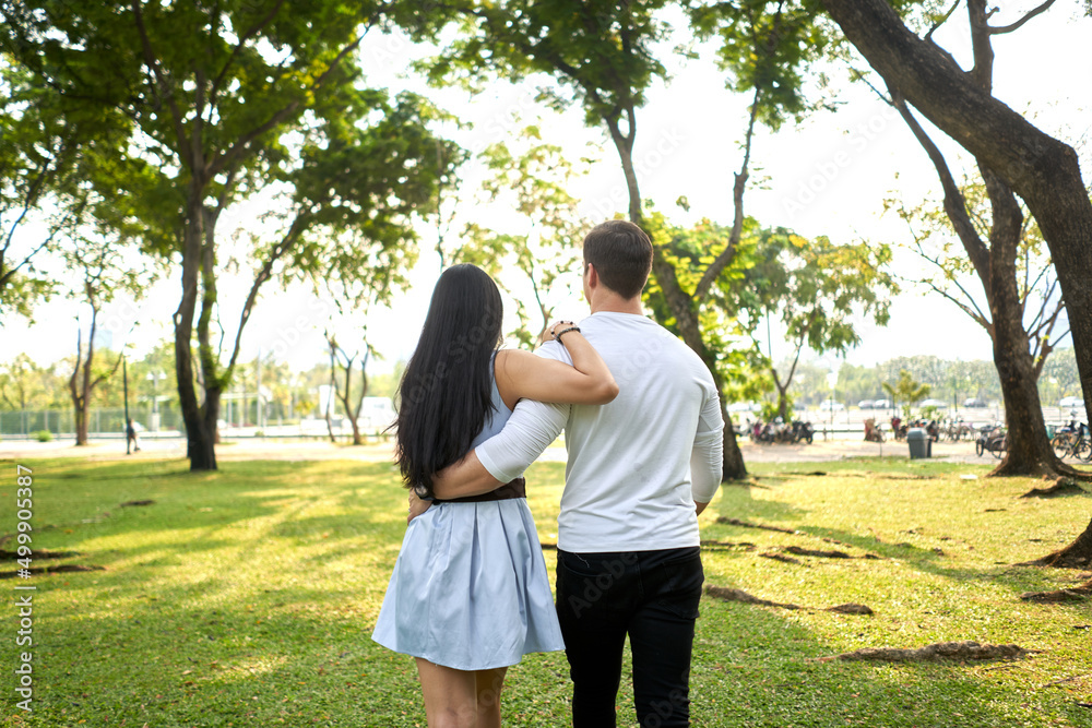 Back of multiracial couple walking in an urban park holding each other's backs Back of multiracial couple walking in an urban park holding each other's backs