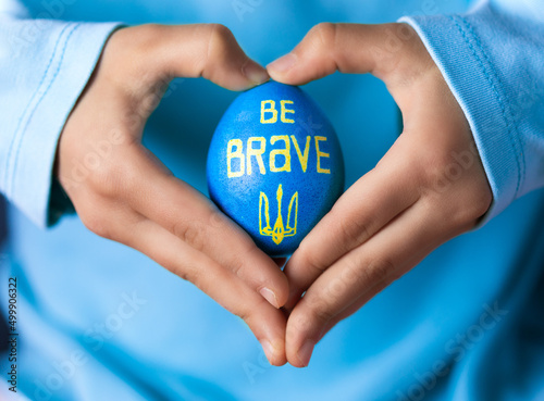 Hands of obscured girl in shape of heart holding Easter egg painted with Ukrainian national colors, blue and yellow, with signature “Be Brave” and Ukrainian national symbol - trident