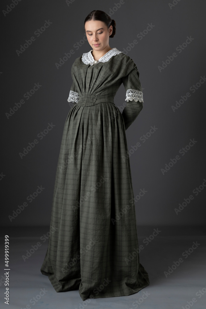 A Victorian working class woman wearing a checked bodice and skirt and standing alone against a studio backdrop