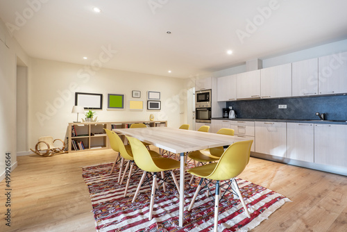 Wooden dining table, yellow resin chairs, wooden kitchen with black wall, wooden shelf and jarapa under the chairs photo