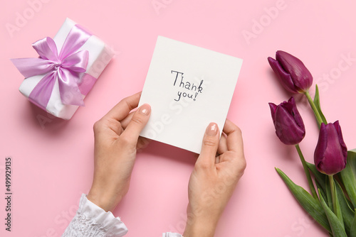 Female hands holding sheet of paper with text THANK YOU, gift box and flowers on pink background