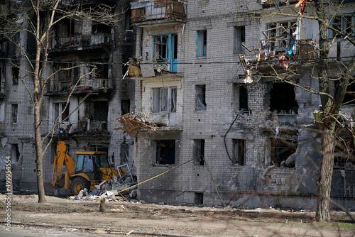 A burnt, destroyed apartment building without windows, damaged during shelling and military operations in Ukraine. Under the house, the tractor clears the area from snares and bricks. © Serhii Prystupa