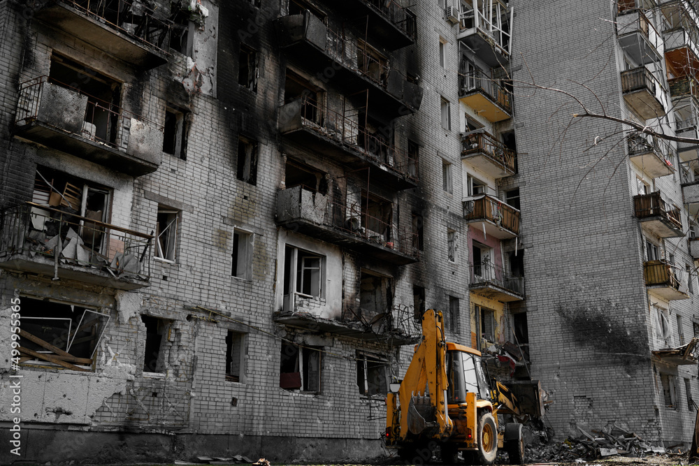 A burnt, destroyed apartment building without windows, damaged during shelling and military operations in Ukraine. Under the house, the tractor clears the area from snares and bricks.