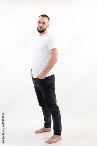 Young caucasian bearded man with short dark hair in white T-shirt, black jeans, standing, thrusting hands into pockets.