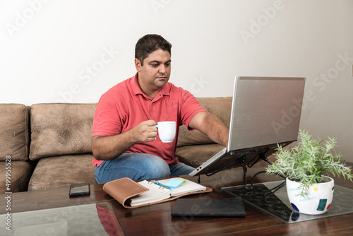 Homem jovem brasileiro em video chamada desde casa com laptop bebendo café photo