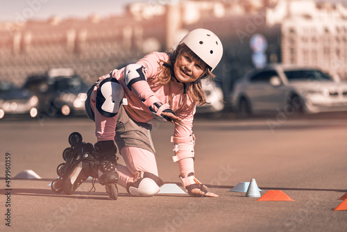 Happy child learns to roller skate on the streets of the city next to the cars. Girl dressed in fall protection trains to ride skates outdoors. The athlete gets up after falling.